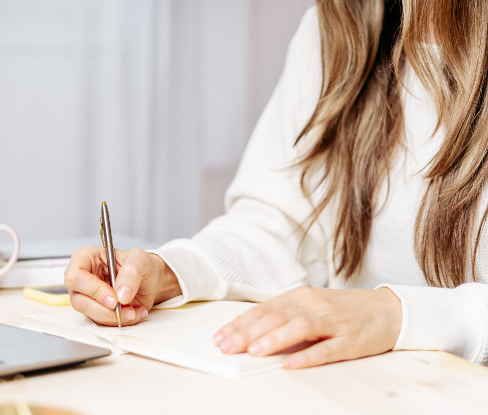 Person writing in a journal with a laptop and coffee cup, symbolizing life planning and self-reflection.