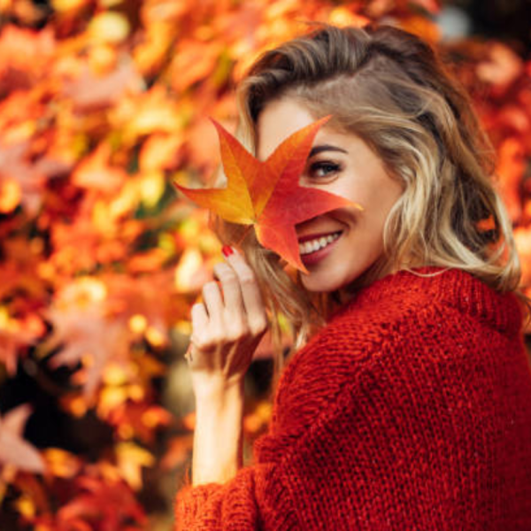Woman smiling while holding a fall leaf, enjoying cozy self-care outdoors in autumn.