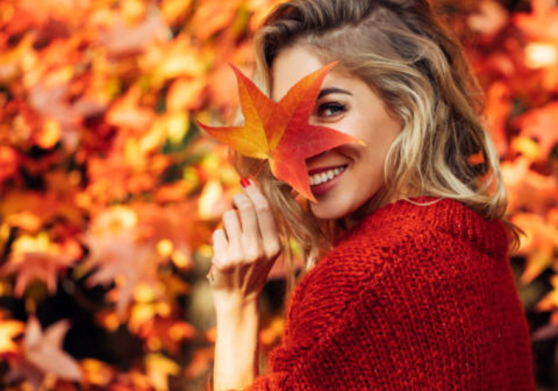 Woman smiling while holding a fall leaf, enjoying cozy self-care outdoors in autumn.