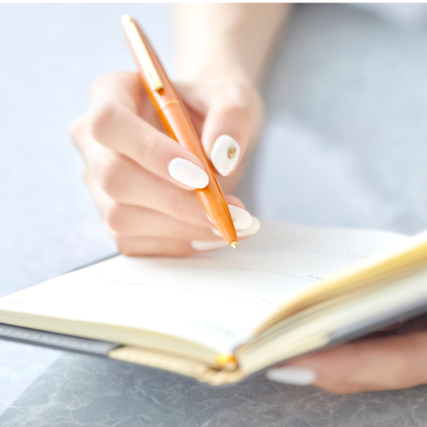 A woman sits at a desk writing in a 2026 planner, surrounded by stationery and a cup of coffee, appearing focused on organizing her schedule.