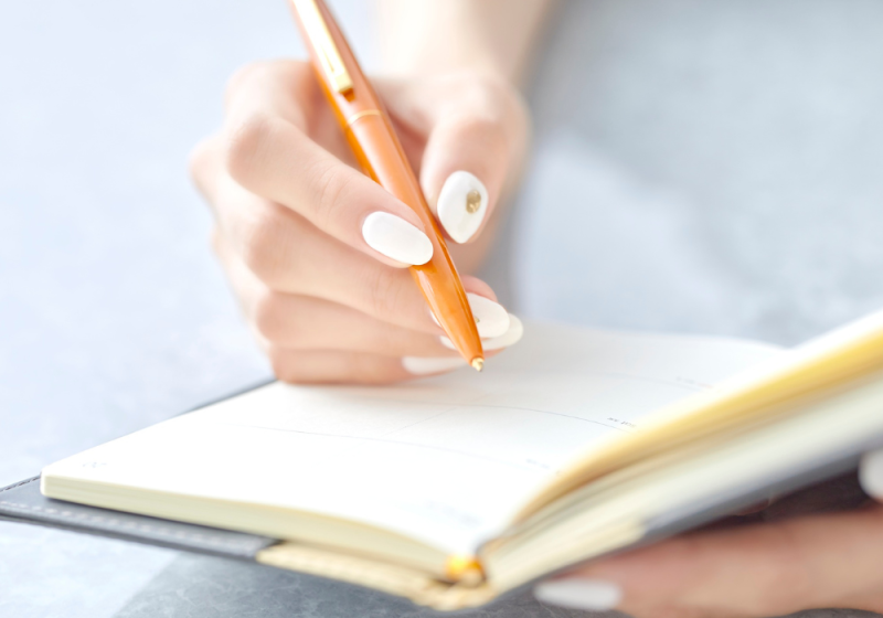 A woman sits at a desk writing in a 2026 planner, surrounded by stationery and a cup of coffee, appearing focused on organizing her schedule.
