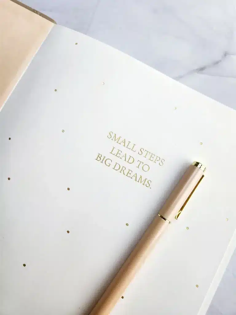 Minimalist white planner cover with gold foil quote βSmall Steps Lead to Big Dreamsβ and blush pink pen on a clean marble background.