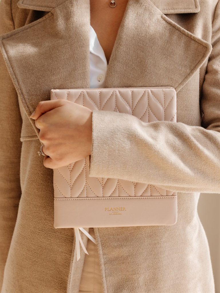 Woman holding a blush pink quilted LH Agenda planner while wearing a beige wool coat, showcasing an elegant luxury weekly planner aesthetic.