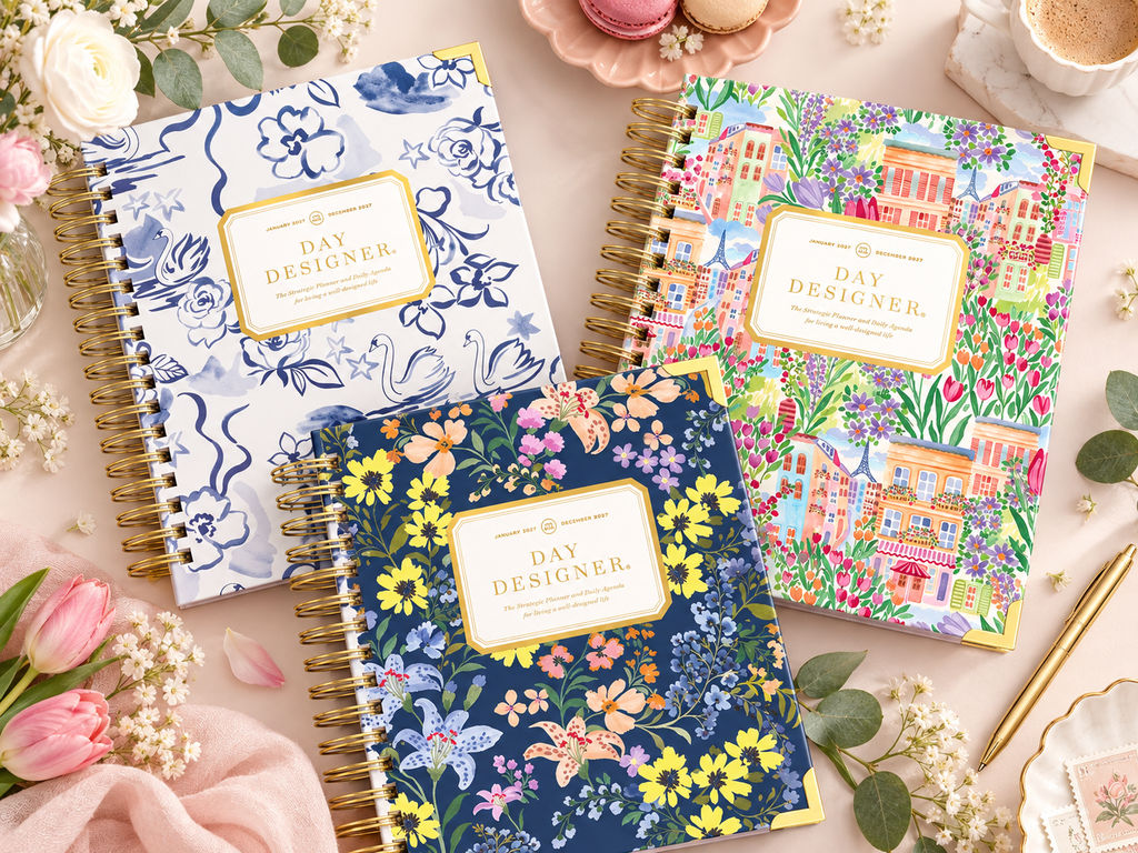 2027 Day Designer planners in blue-and-white swan, colorful Paris floral, and navy floral designs styled together on a feminine desktop with flowers and gold accents.